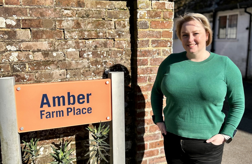 Lisa stands next to a brick wall with an orange sign which says Amber Farm Place in navy text. 