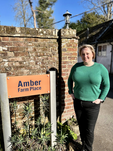 Lisa stands next to a brick wall with an orange sign which says Amber Farm Place in navy text. 